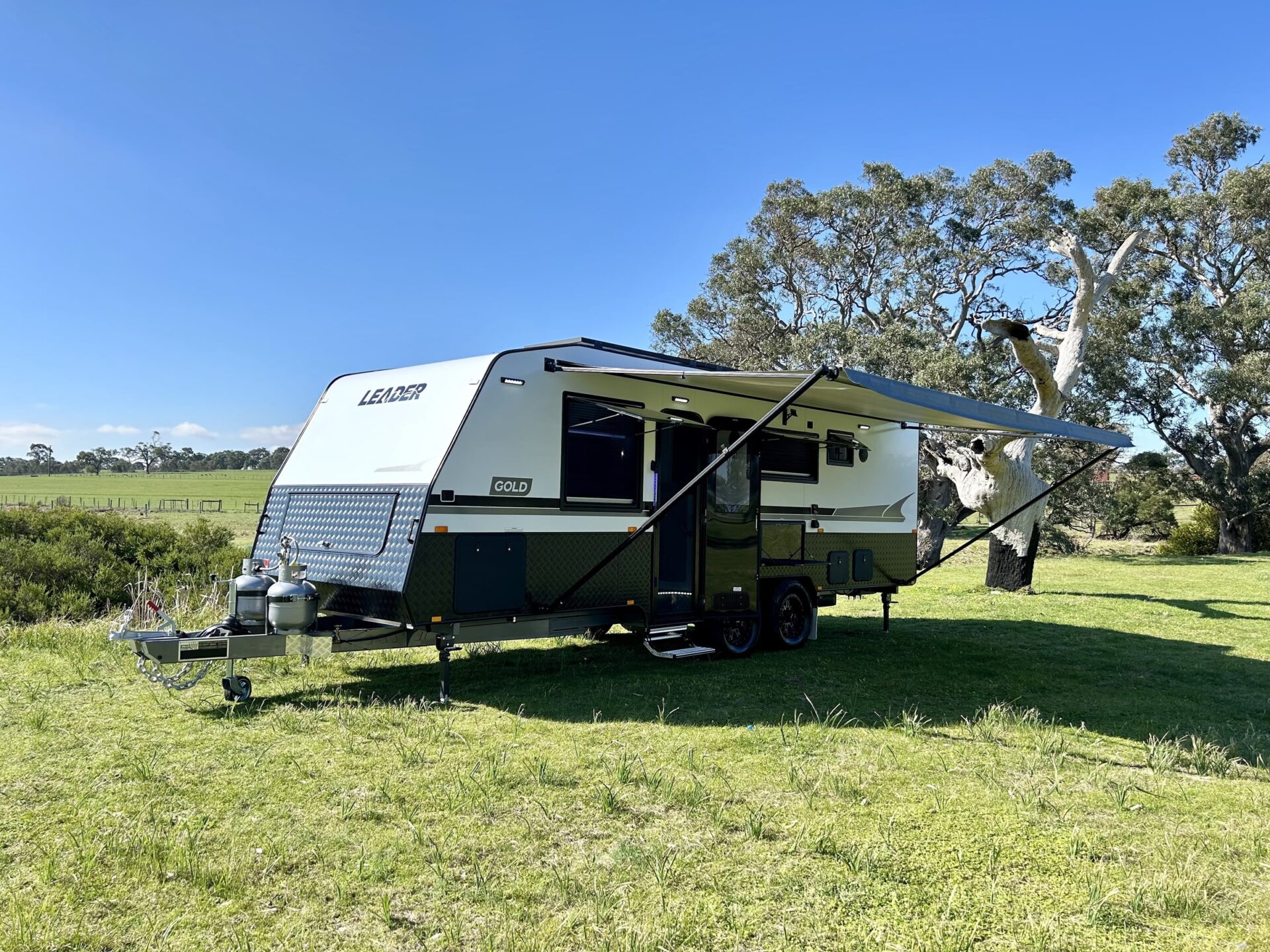 Leader Gold LE caravan exterior — front three-quarter view with twin gas bottles, checker-plate stone guard and tandem axle in a grass paddock