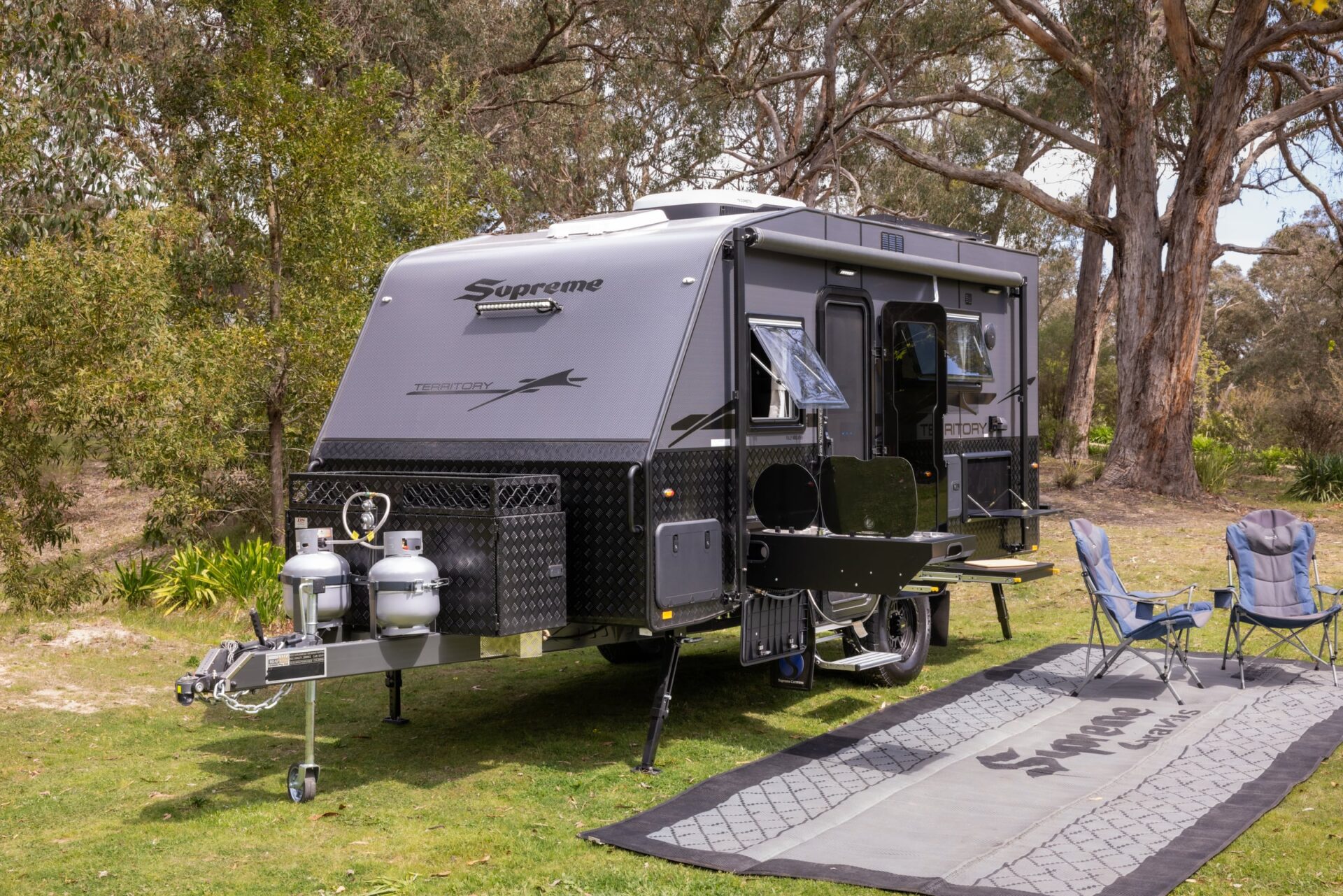 Supreme Territory side profile in bushland — entry door, slide-out kitchen with stove, spare wheel and awning, with camp chairs on Supreme mat