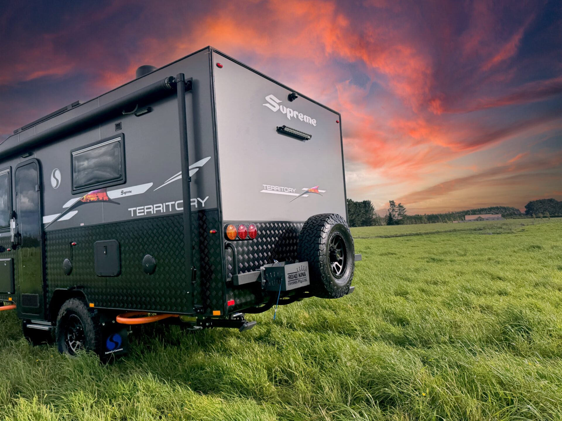 Supreme Territory rear-side angle in a grassy paddock — Territory decals, checker-plate cladding, spare tyre and Road King chassis under sunset sky