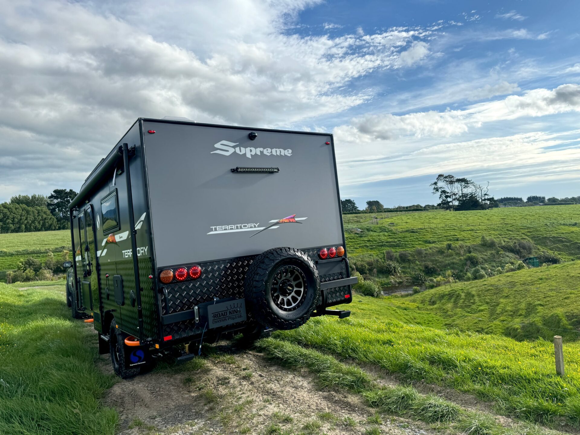 Supreme Territory rear three-quarter on a dirt track — checker-plate armour, spare wheel and tail lights under natural blue sky